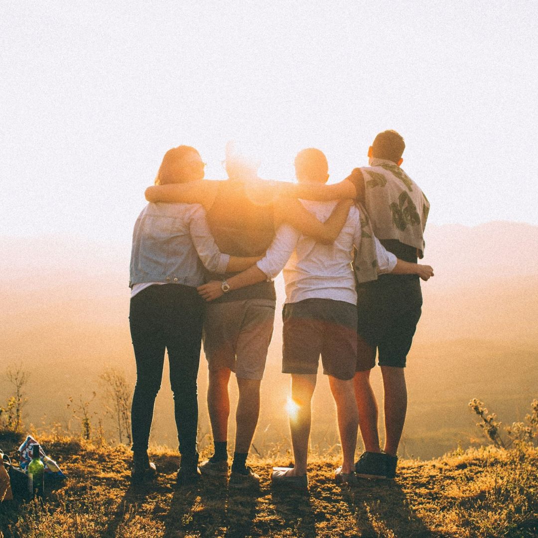 Four young adults standing towards the sunset, with arms around each other's shoulders.